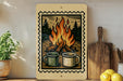 a wooden frame with a printed illustration of two mugs, one green and one white, sitting next to a campfire with flames. The frame is placed on a shelf next to a wooden cutting board and a potted plant.