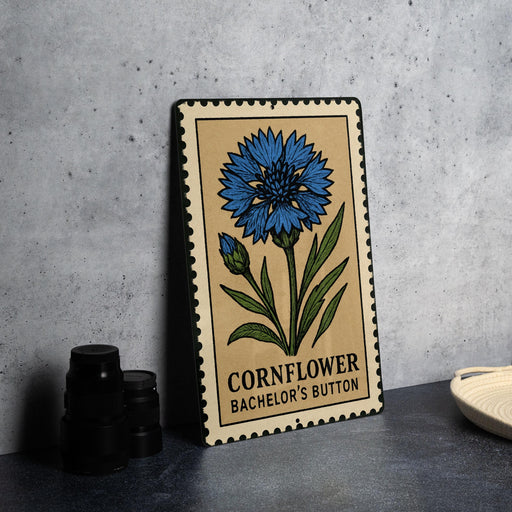 a metal sign with a blue flower illustration and the text "Cornflower BACELOR'S BUTTON" on it, placed on a table next to some black camera lenses.