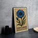 a metal sign with a blue flower illustration and the text "Cornflower BACELOR'S BUTTON" on it, placed on a table next to some black camera lenses.