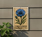 a sign with a blue flower and green leaves, mounted on a wall.