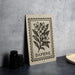 a framed print of a floral illustration with the word "DAPHNE" printed on it, placed on a table next to some black camera lenses.