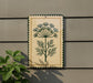 a decorative sign with a plant design on it, mounted on a wall next to a plant.
