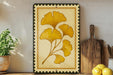 a framed artwork of yellow ginkgo leaves on a wooden shelf, with a potted plant to the left and a wooden cutting board to the right.