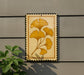 a decorative piece of art with a yellow leaf design, mounted on a wall next to a plant.