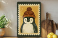 A framed illustration of a penguin wearing a knit hat is displayed on a shelf, accompanied by a wooden cutting board and a potted plant.
