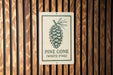 A sign with a pine cone illustration is mounted on a wooden wall.