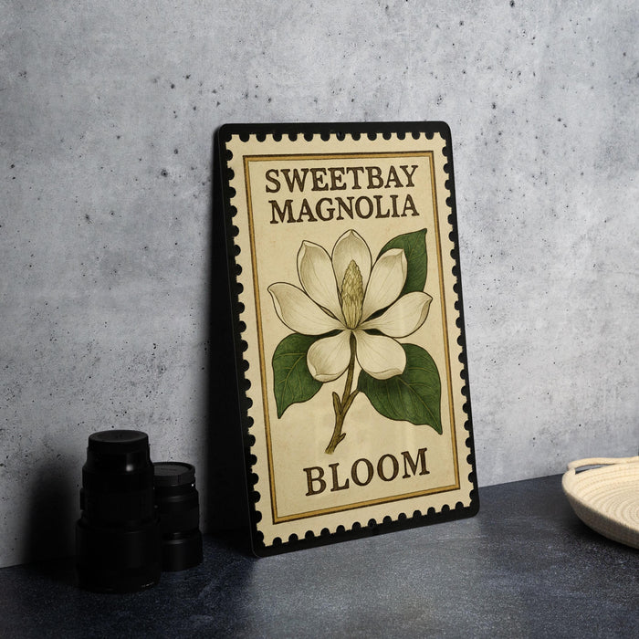 A framed illustration of a white magnolia flower with green leaves, labeled "Sweetbay Magnolia Bloom", is displayed on a gray countertop next to a stack of black camera lenses.