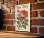 A decorative sign featuring a large red peony flower on a wooden surface against a brick wall background.