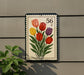 A postage stamp-like sign with a floral design and the number 56 on it, mounted on a wall next to a plant.