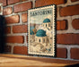 A vintage-style metal sign with a picture of Santorini's iconic buildings, mounted on a brick wall.