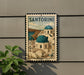 A metal sign with a vintage-style illustration of Santorini, a Greek island, mounted on a wall.