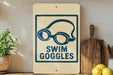 a sign that reads "SWIM GOGGLES" with a graphic of goggles on it, placed on a wooden surface next to a potted plant and a cutting board.