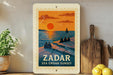 A vintage-style poster of a sunset over the sea, featuring silhouettes of people sitting on a pier. The poster is framed and displayed on a shelf, with a potted plant and a wooden cutting board visible in the background.