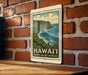 A framed illustration of the Waipio Valley overlooking the ocean, with the text "HAWAII'I WAIPIO VALLEY LOOKOUT" printed on it, is displayed on a brick wall.