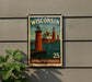A metal sign with a vintage-style illustration of a lighthouse and the text "WISCONSIN" and "LAKESHORE Lighthouses" is displayed on a wall.