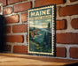 A vintage-style metal sign with the text "MAINE OCEAN SCENIC OVERLOOK" is displayed on a brick wall, depicting a scenic coastal landscape.
