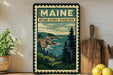 A framed vintage-style poster of Maine's oceanic scenery, featuring a coastal landscape with mountains, pine trees, and the ocean.