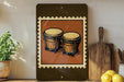 a framed illustration of two wooden drums on a shelf, with a potted plant and a wooden cutting board visible in the background.
