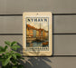 A vintage-style postage stamp depicting a cityscape with colorful buildings and boats, mounted on a wall next to a plant.