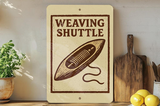 a sign that reads "Weaving Shuttle" with a drawing of a shuttle on it, placed on a shelf next to a wooden cutting board and a potted plant.