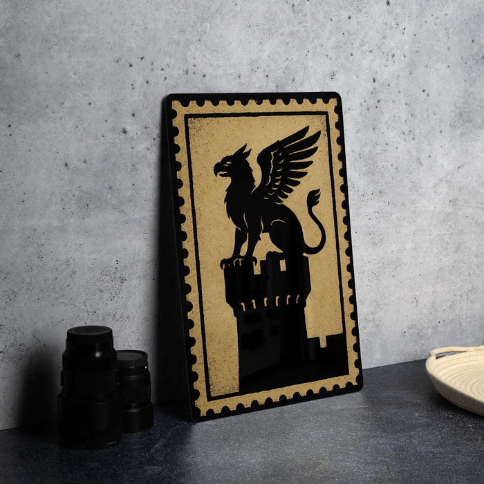 A framed image of a winged lion or mythical creature is displayed on a dark surface, with a stack of black camera lenses and a white dish visible to the side.