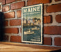 A vintage-style postage stamp featuring a boat illustration and the text "MAINE HISTORIC WATERFRONTS" and "U.S. POSTAGE FOREVER" is displayed on a brick wall.