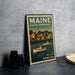 A vintage Maine postage stamp featuring a harbor scene with boats and buildings, placed on a countertop next to some black camera lenses.