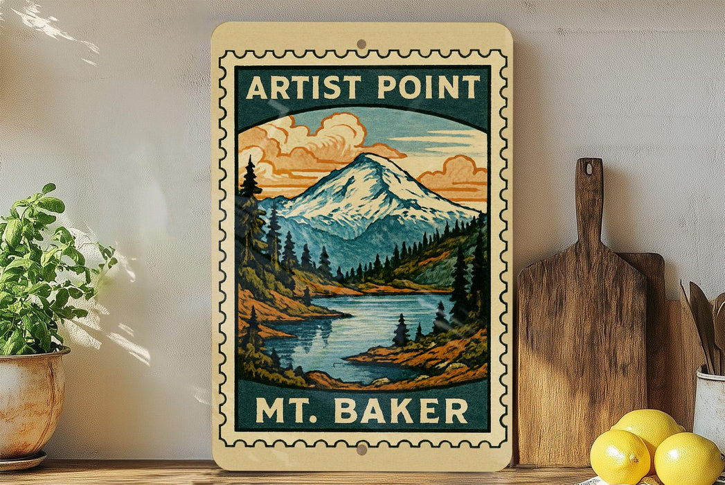 A vintage-style wooden sign with a mountain landscape and a river, featuring the text "ARTIST POINT" and "MT. BAKER". The sign is displayed on a shelf next to a potted plant and a wooden cutting board.