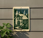 A sign depicting a quaint cottage with a chimney and pine trees, mounted on a wall next to a plant.