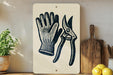 a white sign with a black illustration of a pair of gardening gloves and pruning shears, placed on a wooden surface next to a potted plant and a cutting board.