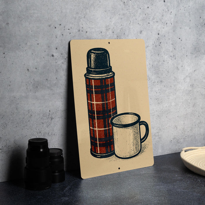A metal sign with an illustration of a thermos and a mug on it, placed on a gray countertop next to some black camera lenses.