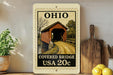 A framed image of a covered bridge in Ohio, with a stamp-like border, is displayed on a shelf next to kitchen utensils and a potted plant.