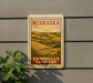 A metal sign with the text "NEBRASKA" and "SANDHILLS COUNTRY" is displayed on a gray wall, with a plant to the left of the sign.