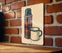 A metal sign with a plaid pattern and a white mug is displayed against a brick wall.