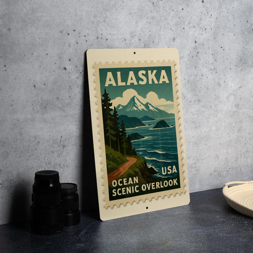 A metal sign with a scenic image of Alaska and the ocean, featuring mountains, trees, and a road, is displayed on a table.
