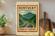 A vintage-style sign for Kentucky Mountain Parkway, featuring a scenic mountain landscape with a winding road, is displayed on a shelf next to a wooden cutting board and a potted plant.