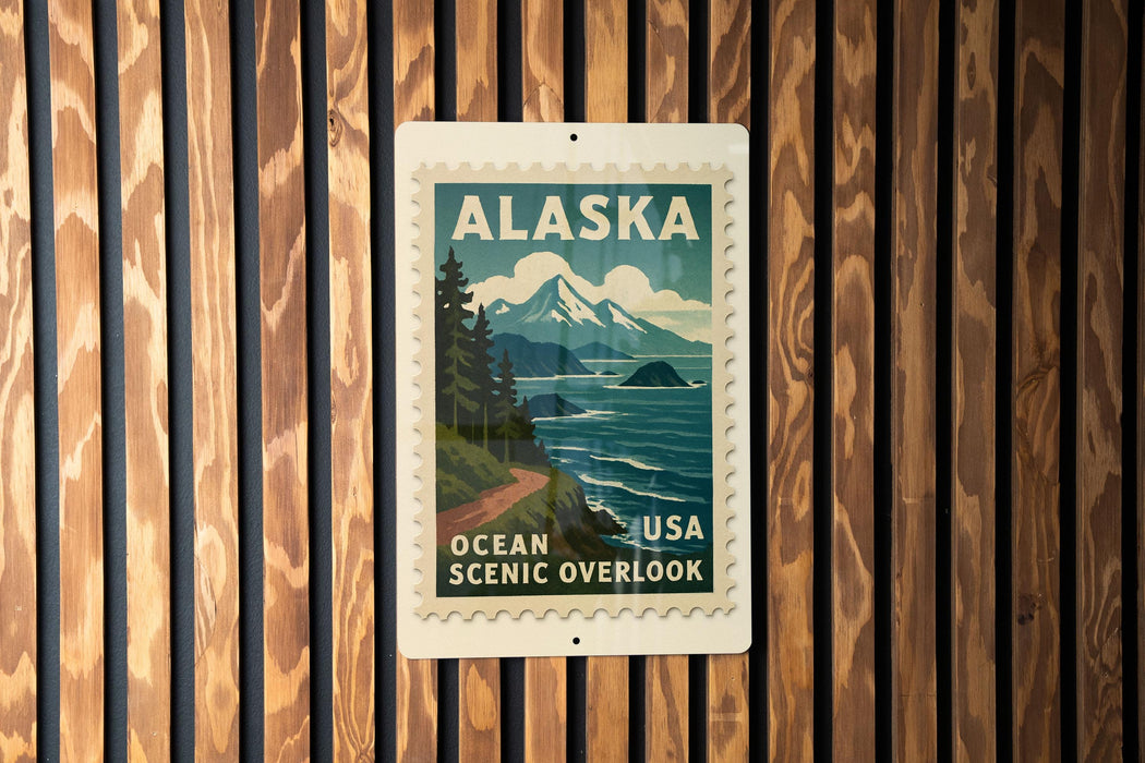 A vintage Alaska postage stamp is displayed on a wooden wall, featuring a scenic mountain landscape with a winding road and ocean in the background.