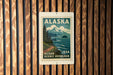 A vintage Alaska postage stamp is displayed on a wooden wall, featuring a scenic mountain landscape with a winding road and ocean in the background.