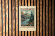 A California postage stamp featuring a scenic landscape with mountains, trees, and a river, mounted on a wooden wall.