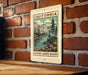 A vintage-style metal sign with a scenic illustration of a mountain landscape and a river, mounted on a brick wall.