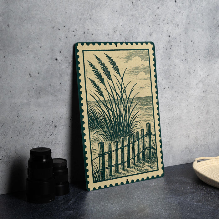 a framed illustration of a beach scene with a wooden fence and grassy plants, placed on a dark surface next to a stack of black camera lenses.