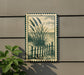 a framed artwork depicting a beach scene with a wooden fence and tall grasses, hanging on a wall next to a plant.