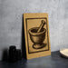 a brown sign with a black illustration of a mortar and pestle on it, placed on a gray countertop next to some black camera lenses.