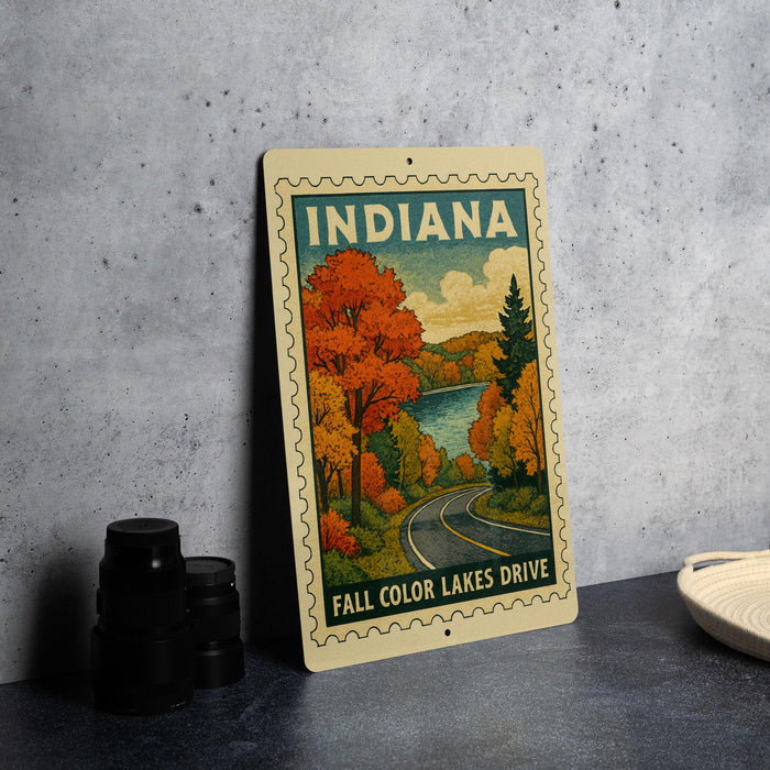 A vintage-style metal sign with the text "INDIANA" and "FALL COLOR LAKES DRIVE" is displayed on a gray concrete surface, accompanied by a stack of black camera lenses and a white dish.