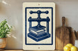 a sign with a blue and white design depicting a book press, placed on a wooden shelf next to a potted plant and a wooden cutting board.