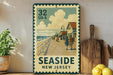A vintage seaside postcard from New Jersey, featuring a wooden pier with people walking along it, is displayed on a wall next to a wooden cutting board and some potted plants.