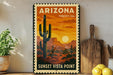 A vintage-style sign for Arizona Forever USA, featuring a desert landscape with a cactus and a sunset, is displayed on a wall next to a wooden cutting board and a potted plant.