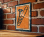 a trombone on a brick wall, with a small orange and blue stamp-like frame surrounding it.