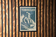 A vintage-style postage stamp featuring a person skiing on a snowy slope, with the word "TOBOGGAN" prominently displayed.