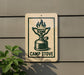 A sign with a graphic of a camp stove is mounted on a wall, with a plant visible to the left.